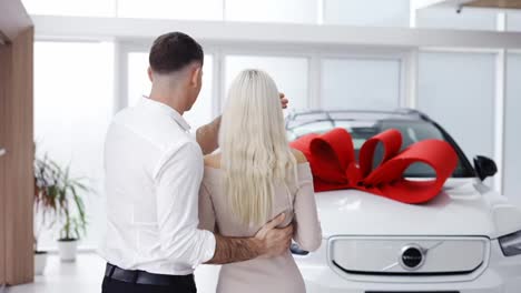 A-man-make-surprise-to-a-blonde-woman,-closed-woman's-eyes-from-back---new-car-with-red-bow,-rear-view