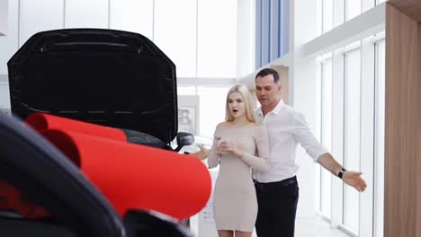A-man-make-surprise-to-a-blonde-woman,-a-new-car-with-red-bow-in-a-showroom