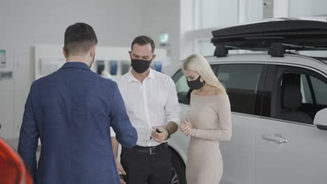 New-car-owners-getting-keys-to-purchased-luxury-auto-in-dealership,-wearing-masks