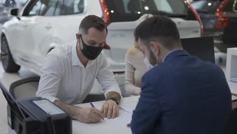 Couple-in-masks-purchase-of-a-new-car,-signing-the-contract-in-the-showroom
