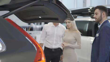 Car-agent-showing-trunk-to-customers-car-in-automotive-dealership-showroom
