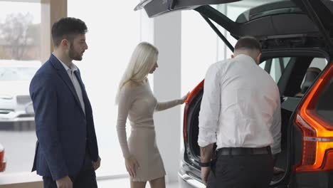 Salesman-showing-the-capacity-of-a-trunk-from-inside-to-customers-in-car-showroom,-side-view