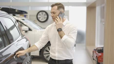 Young-man-using-phone-while-chooses-a-new-model-of-a-luxury-car-in-showroom