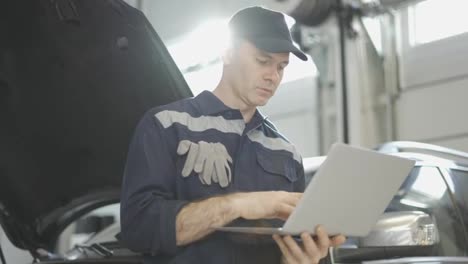 Car-mechanic-working-on-laptop-in-auto-repair-service,-lean-on-a-car,-low-angle-view