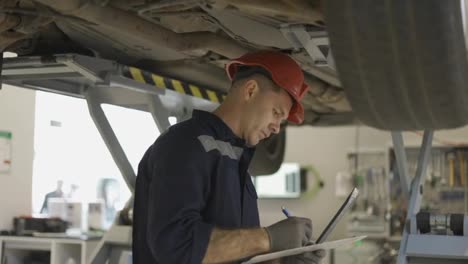 A-car-mechanic-standing-under-lifted-car,-making-notes-on-a-tablet