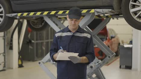 A-car-mechanic-standing-next-to-lifted-car,-making-notes-on-a-tablet