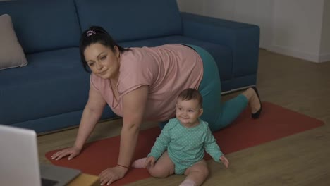 Overweighted-woman-doing-exercises-online-from-laptop-with-little-baby-next-to-her