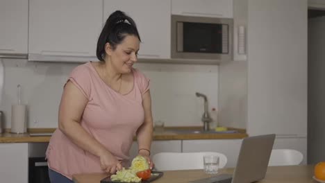 Young-overweight-woman-cooking-salad-at-kitchen,-watching-video-recipe-on-laptop-online