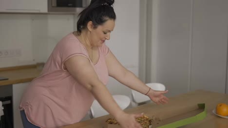 Plus-size-girl-opening-box-with-pizza-inside-in-the-kitchen-and-enjoying-it
