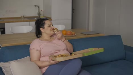 Plus-size-woman-eats-slice-of-pizza-with-great-pleasure-sitting-on-a-couch