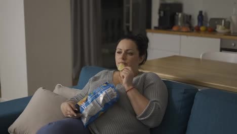 Overweight-woman-sitting-on-couch-at-home,-watching-tv-and-eating-potato-chips