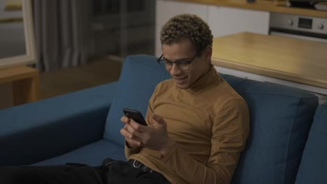 Curly-young-man-web-surfing-on-his-mobile-phone-on-the-sofa-at-home