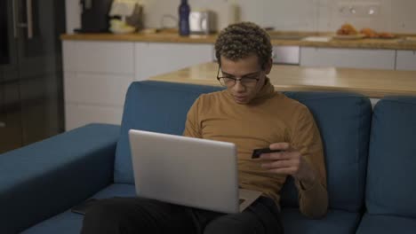 Young-man-buying-online-uses-a-laptop-and-a-credit-card-on-sofa-home