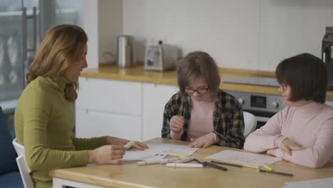 Two-girls-with-disabilities-draw-at-the-kitchen-next-to-the-teacher-or-mother