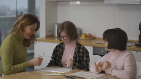 Two-girls-with-syndrome-draw-at-the-kitchen-next-to-the-teacher-or-mother-together,-slow-motion