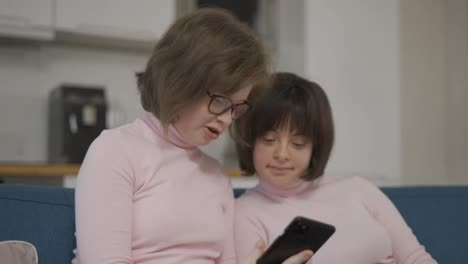 Zwei-Mädchen-Mit-Down-Syndrom-Schauen-Gemeinsam-Auf-Einer-Couch-Auf-Den-Bildschirm-Ihres-Smartphones