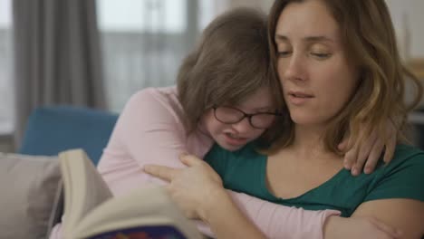 Young-mother-reading-a-book-to-her-daughter-with-down-syndrome-while-they-sitting-on-sofa-in-the-room