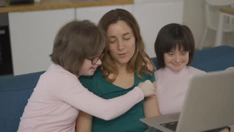 Mother-with-two-daughters-down-syndrome-sitting-on-sofa-have-fun-using-laptop,-slow-motion