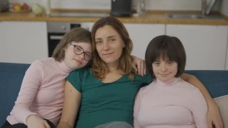 Portrait-of-two-girls-with-down-syndrome-in-sitting-on-a-couch-together-embracing-with-their-mom,-close-up