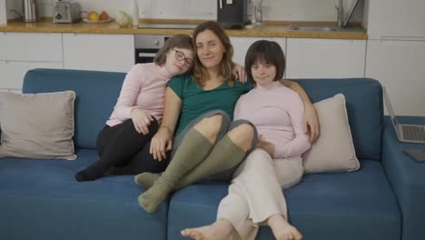 Portrait-of-two-girls-with-down-syndrome-sitting-on-a-couch-together-embracing-with-their-mom