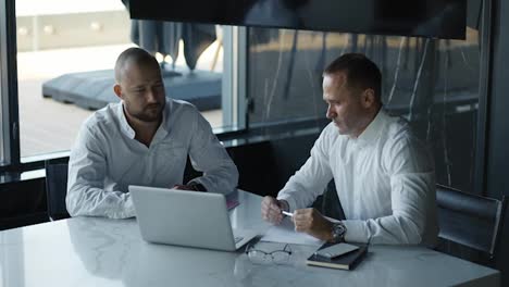 Two-diverse-businessmen-discussing-financial-market-data-using-laptop-and-digital-tablet.-Financial-advisor-broker-manager