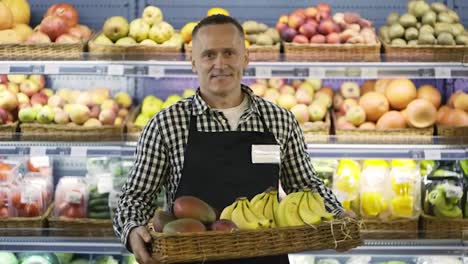 Portrait-of-a-happy-middle-aged-store-worker-holding-a-box-of-exotic-fruits.-A-man-in-an-apron-and-with-a-badge.-Shop-working