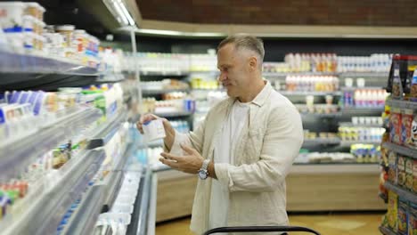 A-middle-aged-visitor-in-a-grocery-store-chooses-dairy-products.-A-man-with-a-cart-in-his-hands-walks-through-the-store.-Grocery-store-shopping-concept