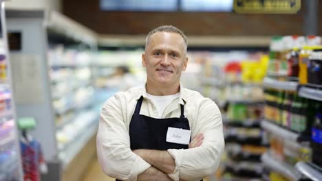 Portrait-of-a-middle-aged-shop-worker-in-an-apron.-The-man-puts-his-hands-on-his-chest.-The-concept-of-working-in-a-grocery-store