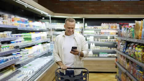 A-happy-middle-aged-shot-visitor-walks-along-the-shelves-with-goods-and-smiles-while-looking-at-the-phone.-The-concept-of-happy