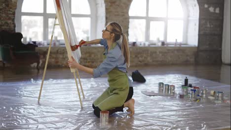 Female-artist-working-with-large-paintbrush-and-wide-canvas-in-workshop-standing-on-knees