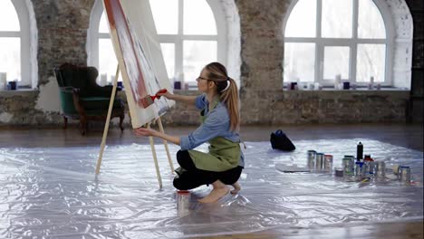 Artist-working-with-large-paintbrush-and-wide-canvas-in-workshop-standing-on-knees
