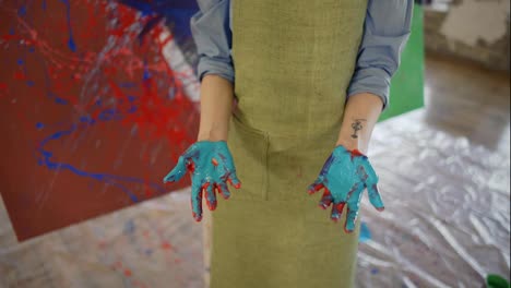 Portrait-of-a-woman-showing-hands-in-paints,-standing-next-to-her-artwork