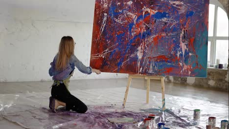 Woman-uses-wide-brush-and-color-paints-splashing-gesture-on-canvas-standing-on-knees