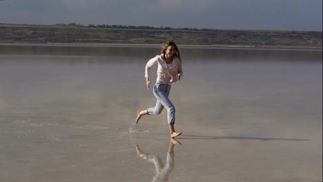 Happy-long-haired-woman-in-casual-is-running-on-sandy-shallow-of-lake-at-summer-and-smiling