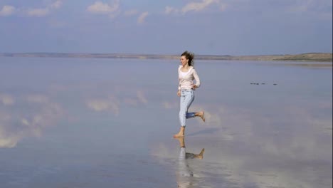 Happy-long-haired-woman-in-casual-is-running-on-sandy-shallow-of-lake-with-mirror-surface-reflection