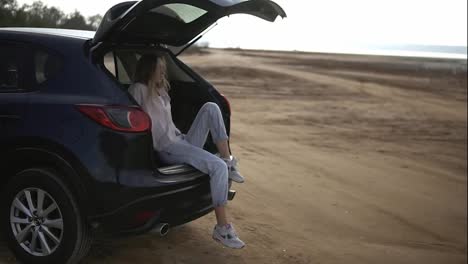 Woman-traveler-having-a-break-sitting-in-car-trunk,-looking-on-waterfront-on-horizon