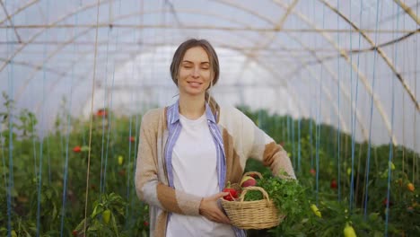 Happy-farmer-woman-holding-basket-with-fresh-harvested-vegetables-and-smiling-at-greenhouse
