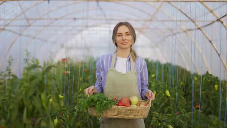 Happy-farmer-woman-walking-with-basket-with-fresh-harvested-vegetables-at-greenhouse