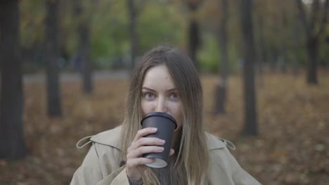 Portrait-of-a-blonde-woman-drinking-take-away-coffee-in-the-golden,-autumn-park
