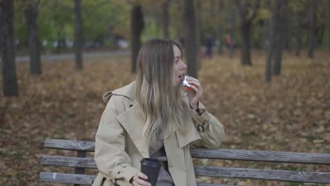 Mujer-Bebiendo-Café-Para-Llevar-Y-Comiendo-Chocolate-En-El-Parque-De-Otoño