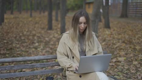Young-beautiful-woman-of-European-appearance-works-at-a-laptop,-sitting-on-a-park-bench