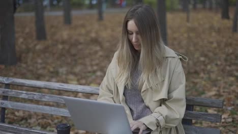 Young-beautiful-woman-works-at-a-laptop,-sitting-on-a-park-bench-with-coffee