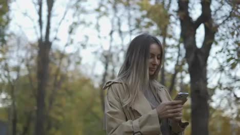 Smiling-woman-reading-messages-or-surfing-internet-from-phone-at-autumn-park,-overview-footage