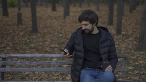 Brunette-man-sits-on-bench-in-the-autumn-park,-works-on-the-phone