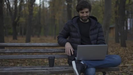 Smiling-man-sitting-casually-on-bench-in-public-park,-using-laptop-for-video-call