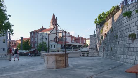 A-well-on-the-Five-Wells-square-and-pedestrians,-with-church-tower-in-the-background
