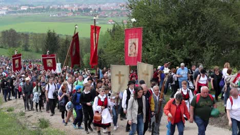 Pilgrims-from-Csíkzsögöd-walk-dirt-road,-carrying-flags-crucifix
