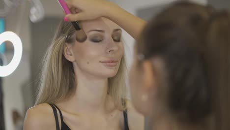 The-make-up-artist-applies-the-powder-on-the-surface-of-the-young-blonde's-face,-slow-mo