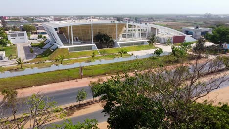 Descending-drone-shot-of-the-Gambia-National-Assembly-building-overlooking-the-busy-road-with-trees-in-the-foreground-on-a-sunny-summer-day