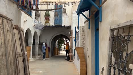 FPV-walking-along-Houmt-Souk-market-meanders-while-a-woman-takes-picture-of-man,-Djerba-island-in-Tunisia